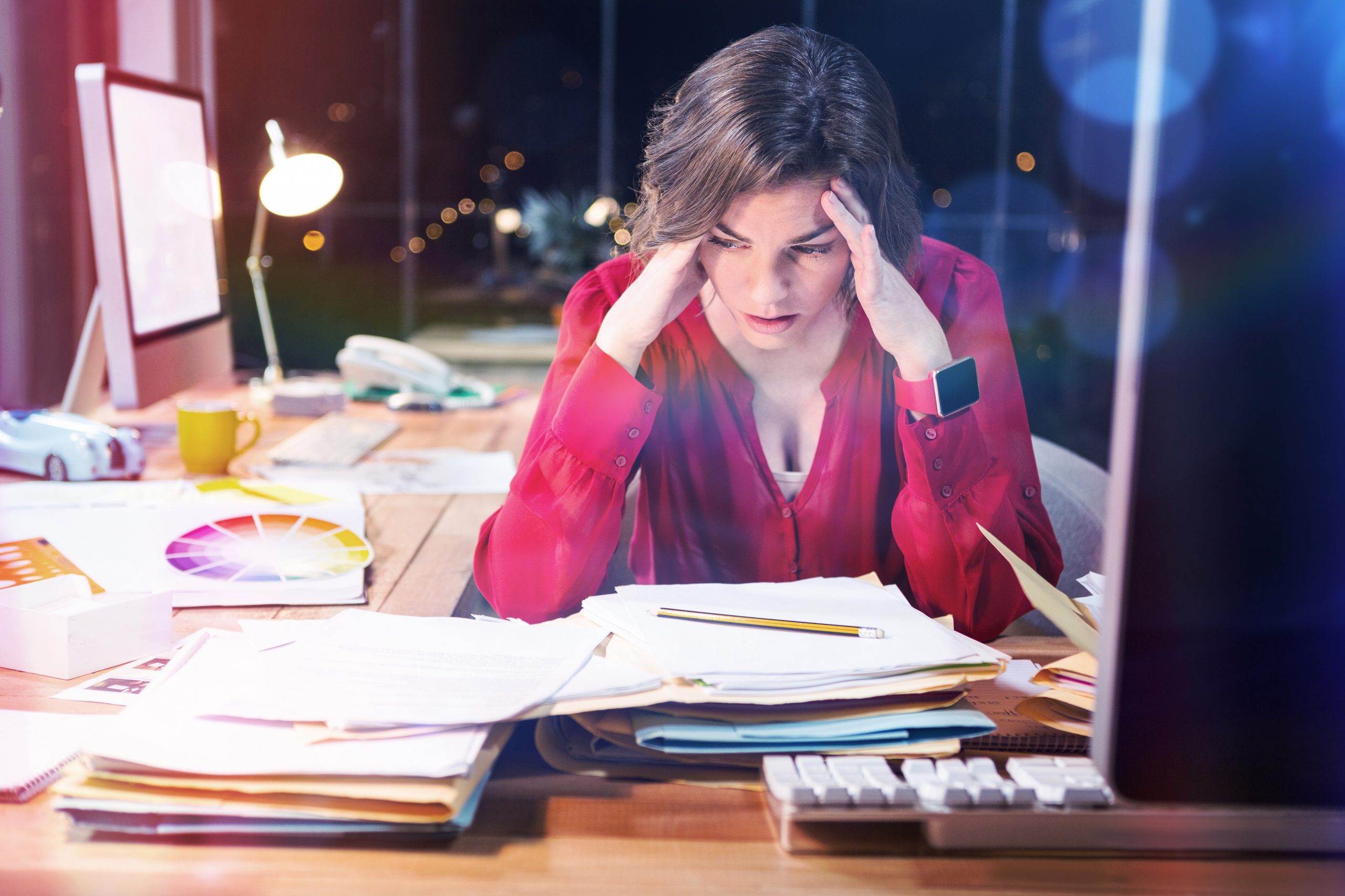 Wypalenie zawodowe - zmęczona, zestresowana kobieta trzymająca się za głowę siedząca przy biurku pełnym papierów